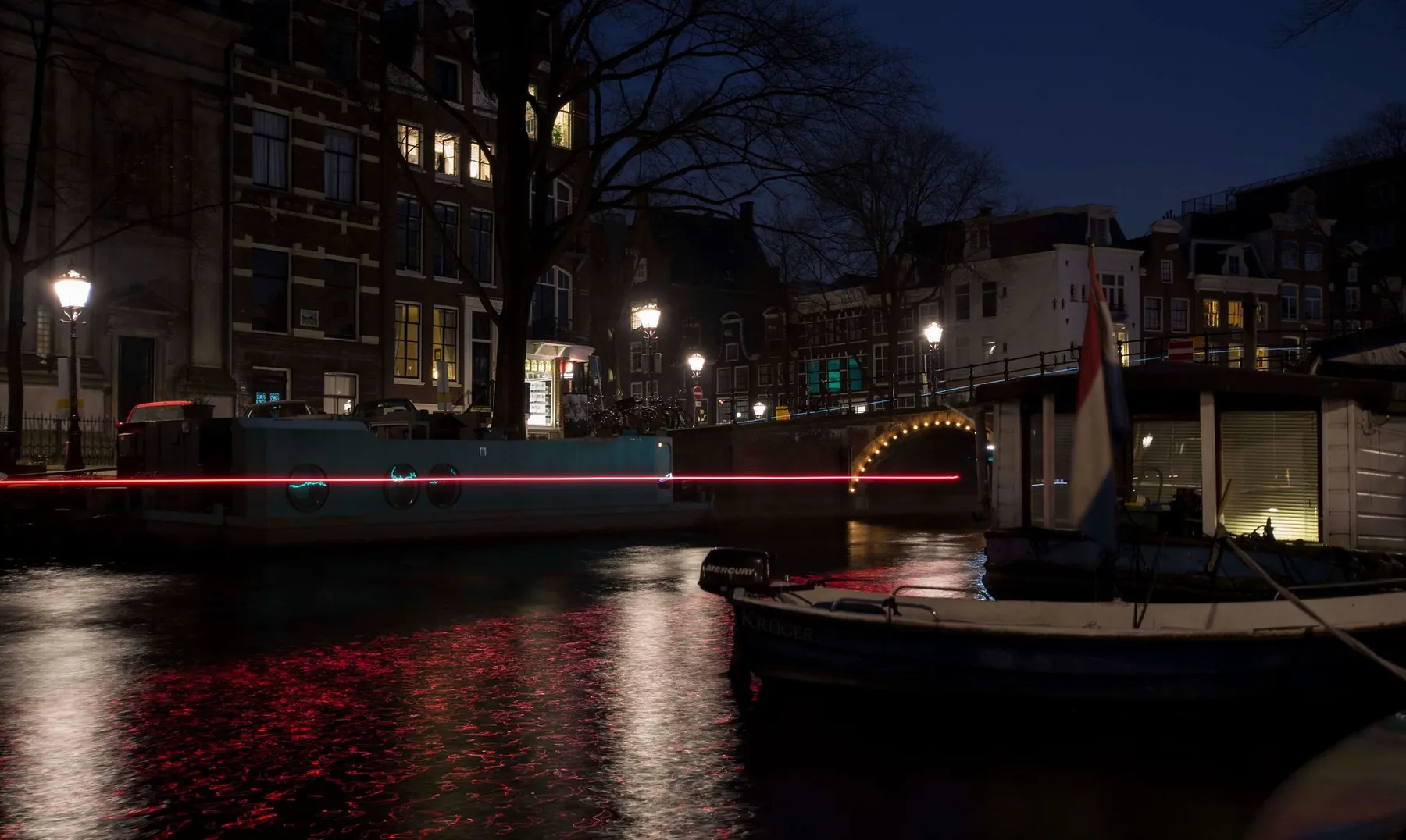 Een Amsterdamse gracht op het blauwe uur, met het rode lichtspoor van een passerende boot voor een brug