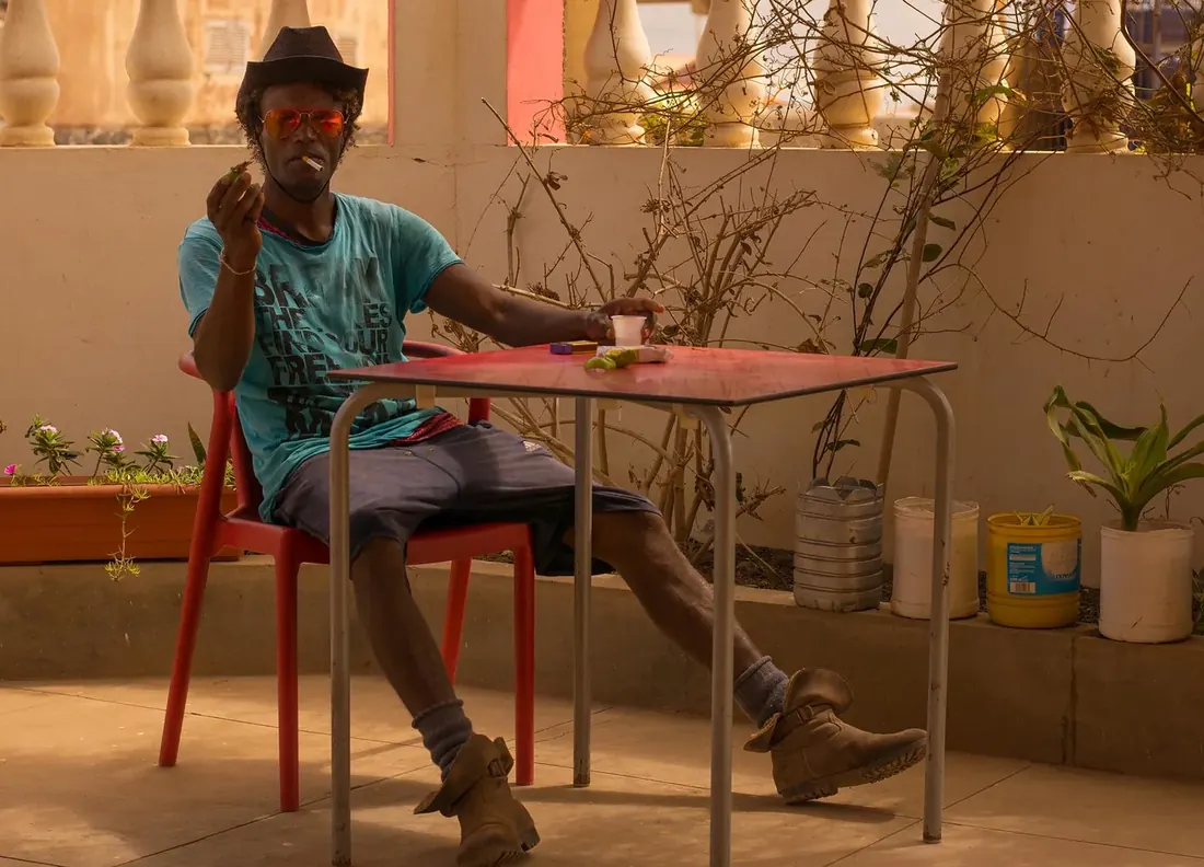 Man in a hat and orange sunglasses at a red table, smoking