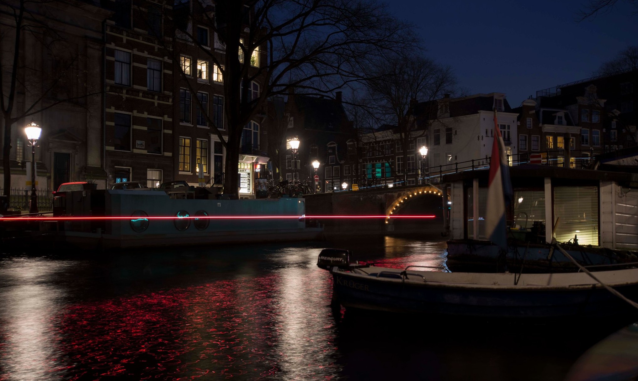 An Amsterdam canal at blue hour, a red light trail from a passing boat crossing in front of a bridge